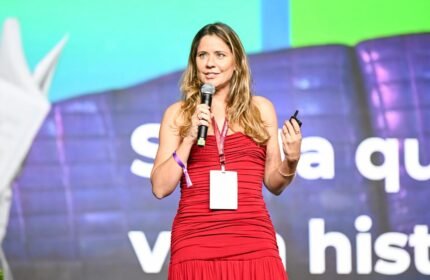 Psicóloga Mayra Gaiato de vestido vermelho segurando microfone durante palestra sobre autismo